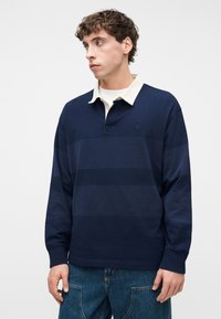 Νεαρός άνδρας που φοράει μπλε ρούκι μπλούζα με σκούρες μπλε ρίγες και λευκό γιακά, συνδυασμένη με μπλε τζιν τζην, κοιτάζοντας προς τα αριστερά.