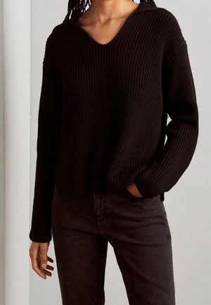Schwarzer, gestrickter Pullover mit V-Ausschnitt und gerippter Textur, mit langen Ärmeln und lockerer Passform. Kombiniert mit dunkel gefärbten Hosen.