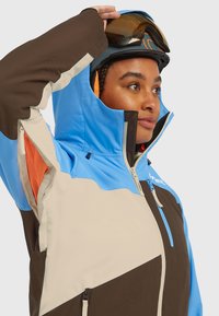 Slēpošanas jaka zilā, brūnā un krēmkrāsā ar augstu apkakli, rāvējslēdzēja kabatām un tīkliņa oderi. Aprīkota ar kapuci un regulējamām aprocēm.