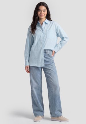 Junge Frau mit langen dunklen Haaren, die ein hellblaues Hemd mit Knöpfen, weit geschnittene Jeans und beige Sneakers trägt, steht mit einer Hand in der Hüfte.