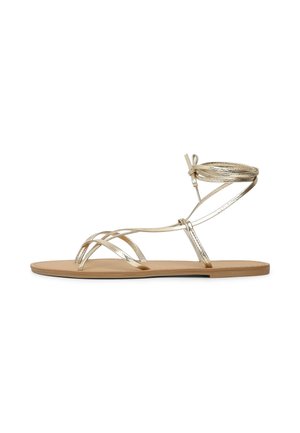 Sandale plate avec fines brides métalliques dorées, comprenant des boucles pour les orteils et un lien en tour de cheville sur une semelle beige.