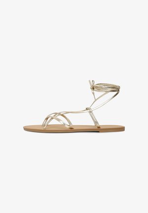 Sandale plate avec fines brides métalliques dorées, comprenant des boucles pour les orteils et un lien en tour de cheville sur une semelle beige.