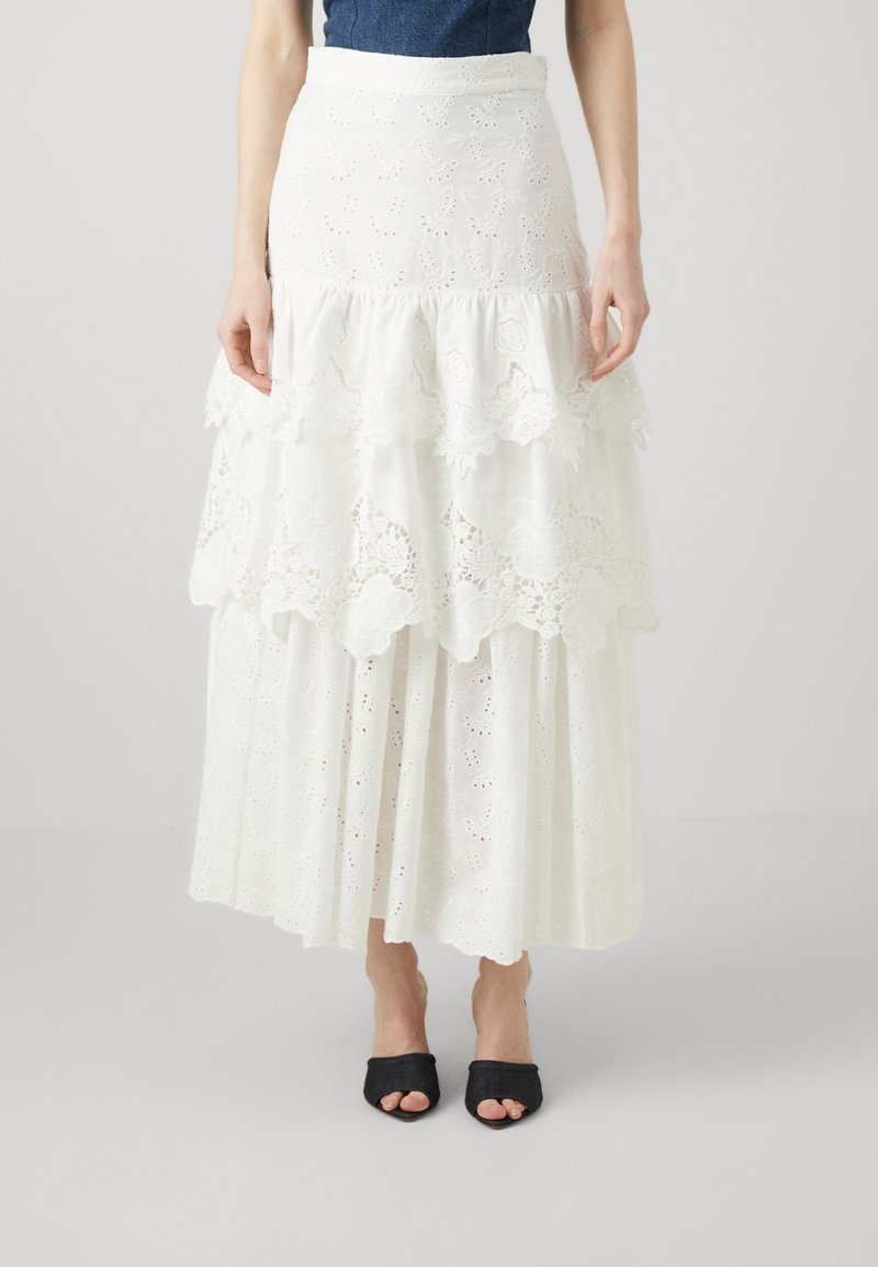 Falda blanca en capas hecha de algodón bordado, con detalles de encaje, una cintura ajustada y una silueta fluida. Combinada con sandalias negras de punta abierta.