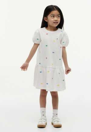 Ragazza giovane con lunghi capelli neri che indossa un vestito bianco con motivi a quadrati colorati, calzini bianchi e sneakers bianche su uno sfondo neutro.