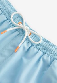 I pantaloni da mare azzurro chiaro presentano una vita elasticizzata con un laccetto bianco, accentuato da punte arancioni. Il tessuto ha una texture liscia.