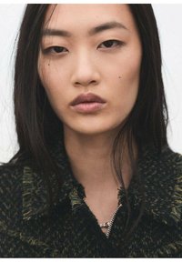 Zbliżenie portretu modelki z długimi, ciemnymi włosami, ubraną w teksturowaną czarno-zieloną kurtkę z kołnierzem, z frędzlami na krawędziach i małym zamkiem błyskawicznym.