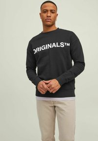 Schwarzes Sweatshirt mit großem weißen "ORIGINALS™"-Schriftzug über der Brust. Gerippte Bündchen und Saum, Rundhalsausschnitt-Design. Getragen mit hellen Hosen.