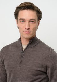Brauner Reißverschluss-Pullover mit strukturiertem Finish, ausgestattet mit einem Stehkragen und einem hellen Hemd darunter. Der Stoff wirkt weich undkörpernah.