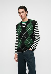 Schwarze Weste mit grünem Rautenmuster und weißen Akzentlinien, übereinander getragen mit einem schwarz-weiß gestreiften Langarmshirt. Dunkle Hose.