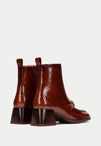 Botas de tobillo de cuero marrón con un acabado brillante, cremallera lateral y un tacón estructurado. Presentan una costura horizontal en la puntera y una pequeña correa decorativa.