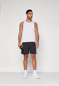 Urheilullinen mies, joka käyttää valkoista hihattomaa toppia ja mustia shortseja, pitää käsipainoa, valkoiset lenkkarit jalassa ja vaatteissa näkyy Puma-logo.