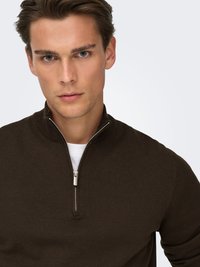 Brown knit pullover with a zip neck design, featuring a textured finish and ribbed cuffs. Worn over a white shirt.