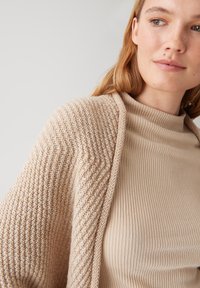 Cardigan beige en tricot avec un motif texturé, porté sur un haut beige en maille côtelée ajusté. Matériau doux et léger, design à ouverture frontale.
