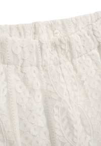 Weißes Spitzengewebe mit einem strukturierten Muster aus kreisförmigen Designs und schmalen vertikalen Streifen; besitzt einen elastischen Bund mit einem Knopfverschluss.