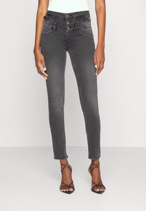 Frau trägt dunkelgraue Slim-Fit-Jeans mit hohem Bund und dreifachem Knopfverschluss sowie schwarze Riemchenabsätze und steht vor einem schlichten Hintergrund.