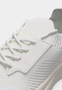 Κοντινό πλάνο ενός λευκού πλεκτού αθλητικού παπουτσιού που δείχνει υφασμάτινη υφή, κορδόνια και λαστιχένιο πλαϊνό πάνελ για υποστήριξη.