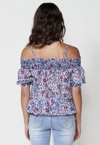 Top floreale a spalla scoperta con scollatura arricciata e maniche corte, caratterizzato da un motivo viola e blu su uno sfondo chiaro.
