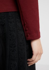 Bordeaux neule pyöreillä hihansuilla, joissa on kolme nappia, yhdistettynä mustaan teksturoituun hameeseen, jossa on monimutkaisia kirjontakuvioita.