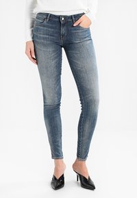 Vaqueros azules de corte slim en denim desgastado con un diseño clásico de cinco bolsillos, cremallera y sutiles detalles de costura. Combinados con tacones negros de punta.