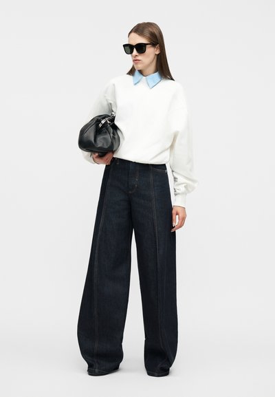Γυναίκα που φοράει μαύρα γυαλιά ηλίου, λευκό πουλόβερ με μπλε γιακά, τζιν με φαρδιά μπατζάκια σκούρου χρώματος, κρατώντας μια μεγάλη μαύρη τσάντα σε απλό φόντο.