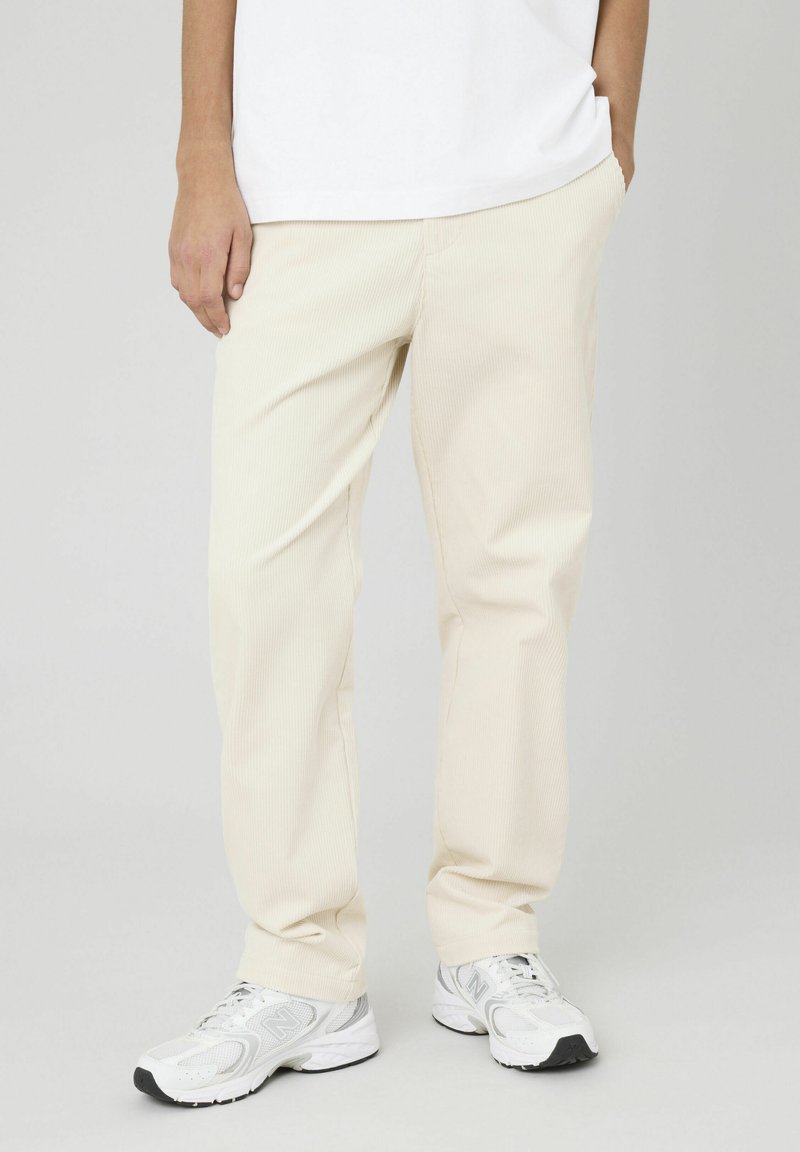 Pantalons en velours côtelé beige clair avec une coupe droite, présentant une texture côtelée subtile et des poches latérales. Associés à des baskets blanches.