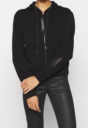 Personne portant un sweat à capuche noir zippé avec une étiquette « guess 1981 » sur la fermeture éclair et un texte sur la manche, associé à un pantalon noir, debout les bras croisés.