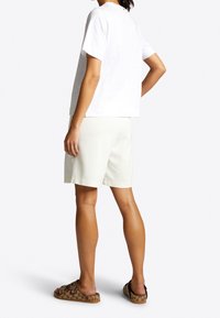 Witte T-shirt met korte mouwen, gecombineerd met lichtgrijze shorts. Het model staat zijwaarts, met bruine sandalen met een patroon. Eenvoudig en casual ontwerp.