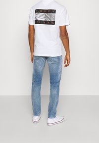 Man med vit Tommy Jeans T-shirt med svart och röd flaggtryck, ljusblå jeans och vita Converse-sneakers, stående mot en vit vägg.