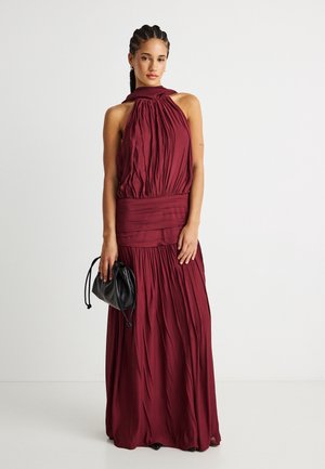 Kvinde med flettet hår iført en ærmeløs, gulvlængde bordeaux kjole, der holder en lille sort pose med snor.