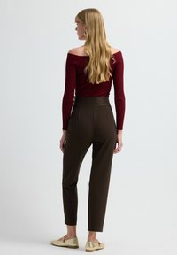 Top burdeos de canalé con escote off-shoulder y mangas largas, combinado con pantalones marrones de cintura alta y zapatos de oro tipo slip-on. Fondo liso.