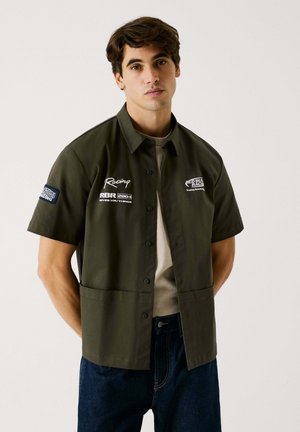Camisa de botones de manga corta, color verde oscuro, con una textura suave; cuenta con logotipos, bordado blanco y un corte relajado. Lleva una camiseta color beige debajo.