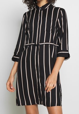 Mujer vestida con un vestido negro abotonado con rayas blancas verticales y mangas tres cuartos, con cinturón en la cintura y cuello.