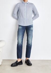 Man som bär en blårutig skjorta, urblekta blå jeans och svarta loafers, stående med händerna bakom ryggen mot en vit vägg.