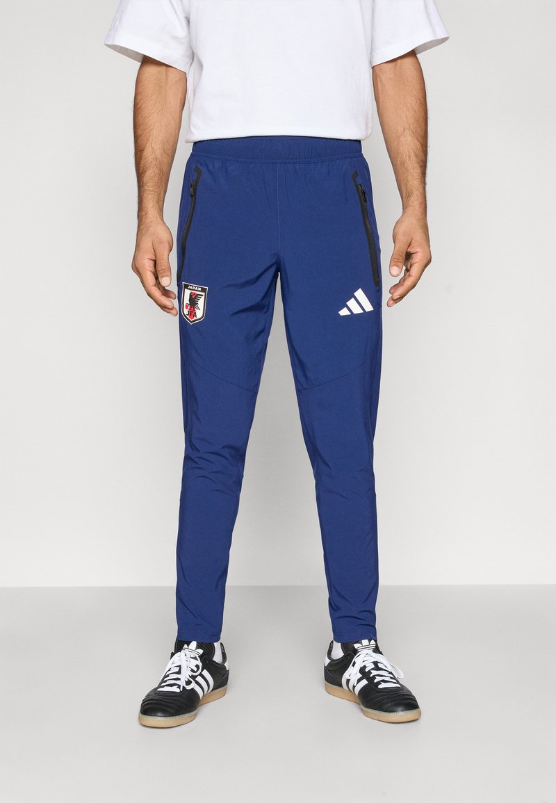 Calças desportivas azuis com cintura elástica, bolsos laterais com fecho e um emblema de logótipo. Feitas de tecido suave, com detalhes contrastantes em preto.