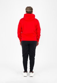 Felpa rossa con cappuccio dalla finitura texturizzata, abbinata a pantaloni della tuta neri a gamba affusolata. Il modello indossa sneakers bianche. Vista da dietro su uno sfondo bianco.