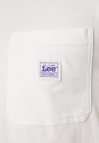 Weißes Baumwoll-T-Shirt mit einer quadratischen Tasche, die mit einem lila gestickten "Lee Workwear"-Label und einem dreieckigen Logo verziert ist. Glatte Textur.