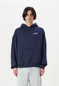 Felpa con cappuccio blu navy, vestibilità rilassata, realizzata in tessuto morbido. Presenta una tasca frontale e un logo bianco sul petto. Polsini e orlo a coste.