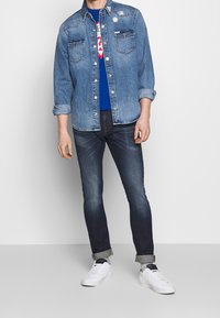 Denim-paita, jossa on rintataskut, sininen t-paita alla, tummat farkut ja valkoiset lenkkarit. Paidassa on nappikiinnitys ja käännetyt hihat.