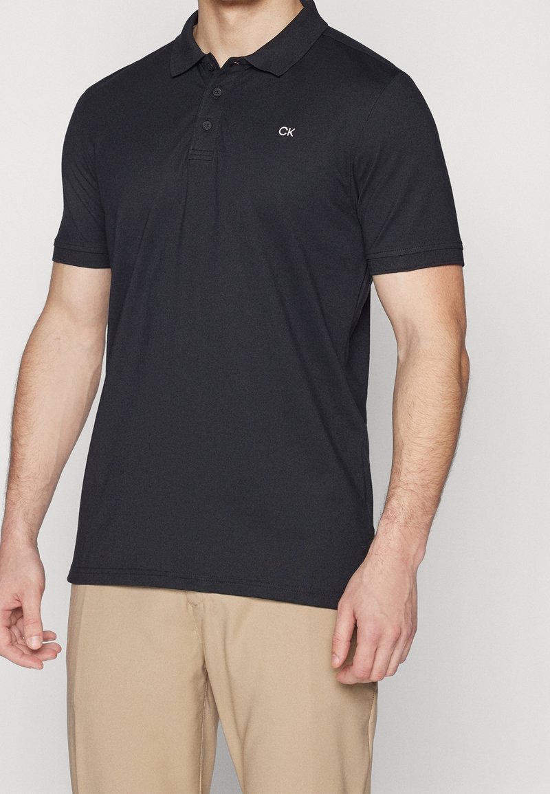 Homme portant un polo noir à manches courtes avec un logo CK sur la poitrine et un pantalon beige, debout devant un fond uni.