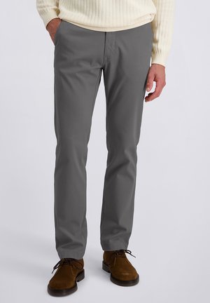 Homme portant un pantalon droit gris, un pull en tricot crème, et des chaussures lacées en suède marron, se tenant avec une main dans la poche.