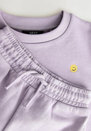 Lilla sweatshirt med ribbet krave, med et gult smiley ansigt broderet. Sweatpants har elastisk talje og snoretr�æk.