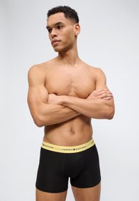 Γυμνό αντρικό πάνω μέρος με μυώδη σωματική διάπλαση που φοράει μαύρα μπόξερ Tommy Hilfiger με κίτρινη ζώνη, με σταυρωμένα τα χέρια, κοιτάζοντας προς τα πλάγια.