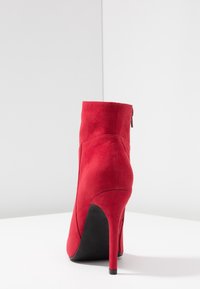 Stivale alla caviglia rosso in suede con tacco alto a punta, caratterizzato da una parte superiore liscia e un retro chiuso con una cerniera laterale. Suola nera visibile.