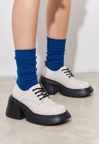 Pantofi cu șireturi, bicolori, cu un exterior lucios de culoare bej și o talpă masivă neagră, asortați cu șosete albastre cu dungi. Textură netedă, formă rotundă.