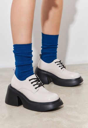 Lacoste, dwukolorowe buty z wiązaniami z błyszczącej beżowej cholewki i czarną masywną podeszwą, w zestawieniu z niebieskimi skarpetami prążkowanymi. Gładka tekstura, zaokrąglony kształt.