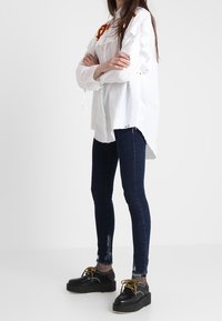 Camisa blanca de gran tamaño con bordado rojo, jeans oscuros ajustados con acentos desgastados y zapatos de plataforma negros con detalles de cordones.