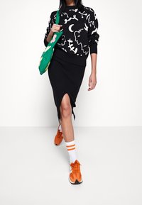 Schwarzer Pulli mit Muster, gerippter schwarzer Rock mit Schlitz an der Seite, orangefarbene Sneaker und eine grüne Tasche mit floralen Motiven. Weiß-orange gestreifte Socken.