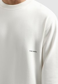 Witte sweatshirt met een geribbelde ronde hals, een gladde textuur en een klein zwart "PURE PATH" logo op de voorkant.