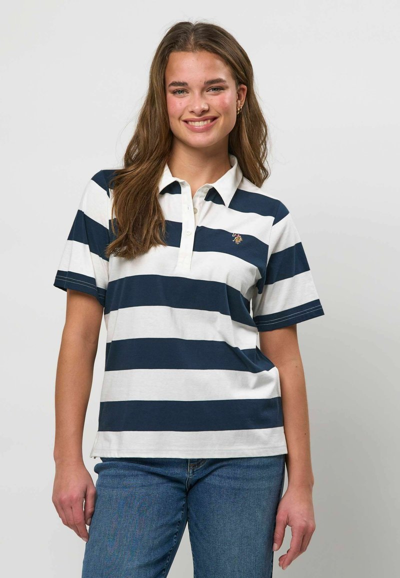 Jeune femme souriante, portant un polo à manches courtes rayé bleu marine et blanc, ainsi qu'un jean bleu, debout devant un fond uni.