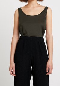 Top in verde oliva con scollatura a barca, abbinato a pantaloni neri a vita alta con texture, caratterizzati da dettagli plissettati.
